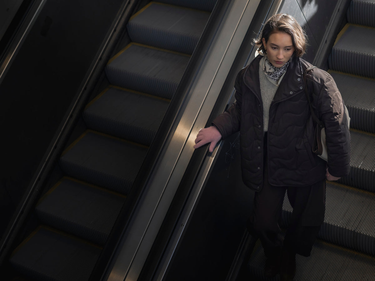Pink knitted gloves worn by a woman in a dark quilted jacket on an escalator in an urban setting.