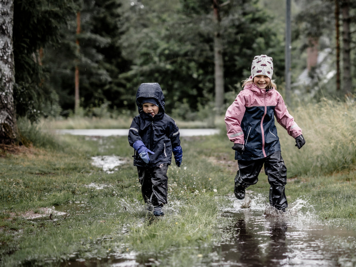 Two children wearing waterproof gloves and rain gear running through puddles on a forest path.